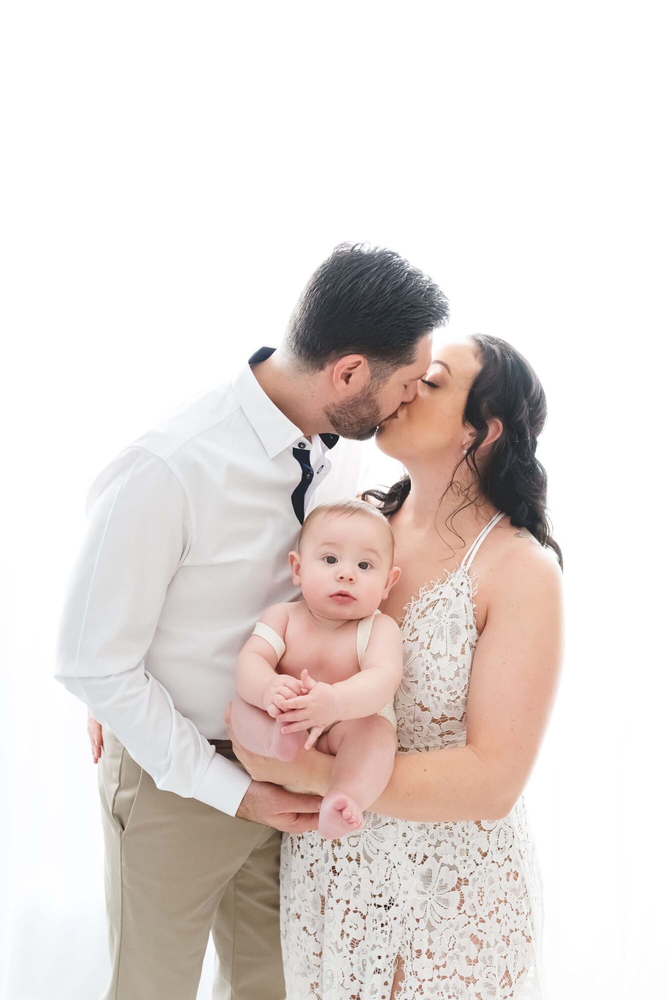 Reaj Roberts Photography Scottsdale family photographer baby held between parents as they kiss in front of a window
