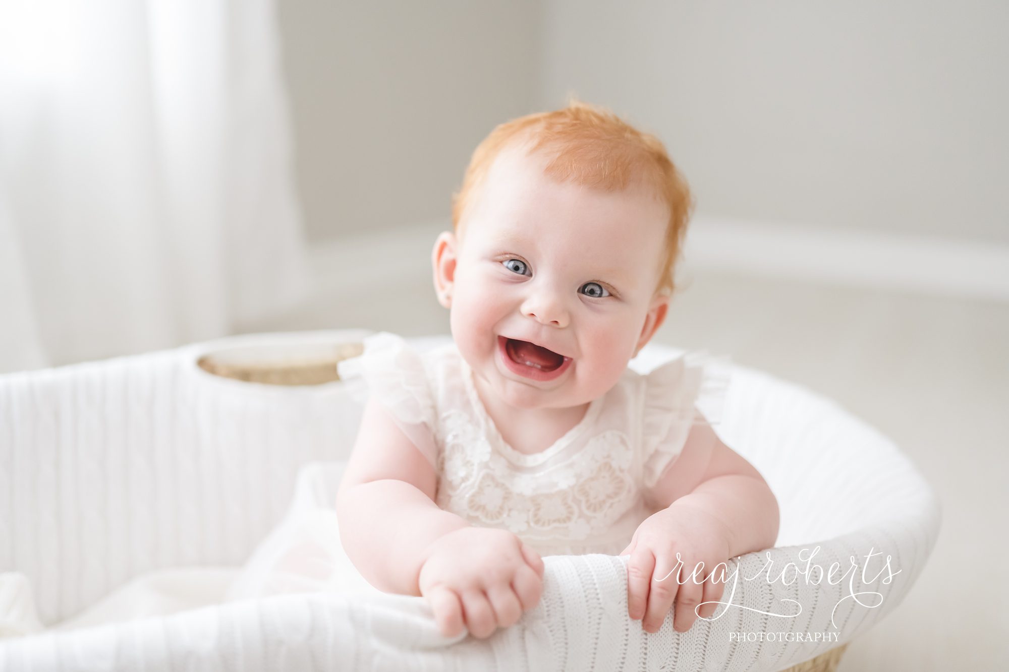 sitter session baby with blue eyes red hair in white dress smiling sitting in moses basket © Reaj Roberts Photography